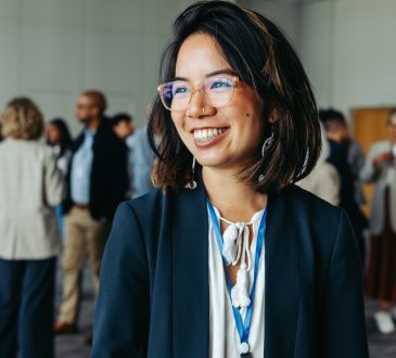 Asian businesswoman in glasses, smiling and networking at a professional conference. Colleagues in the background enhance the atmosphere of collaboration and connection.