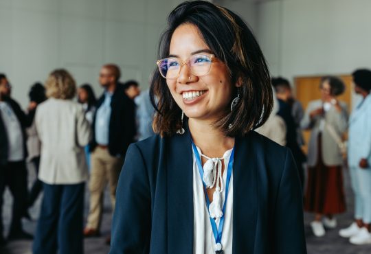 Asian businesswoman in glasses, smiling and networking at a professional conference. Colleagues in the background enhance the atmosphere of collaboration and connection.