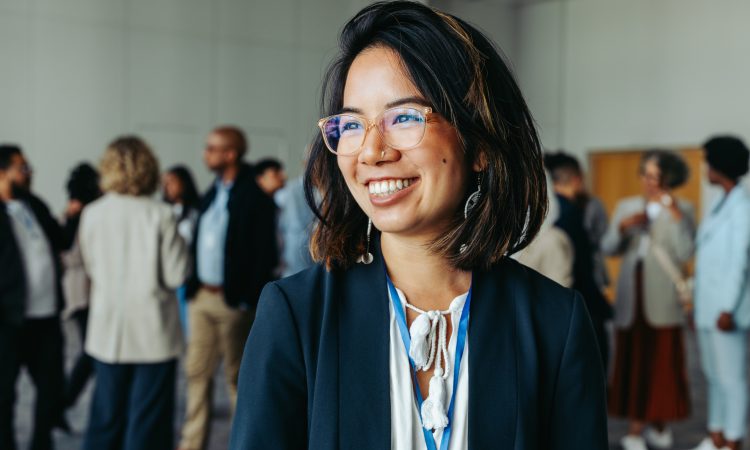 Asian businesswoman in glasses, smiling and networking at a professional conference. Colleagues in the background enhance the atmosphere of collaboration and connection.
