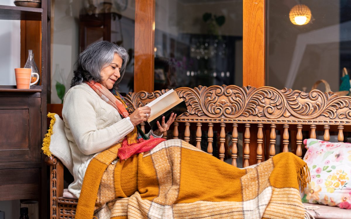 Cozy scene of a senior woman relaxing on a wooden bench with a blanket, reading a book and enjoying the comfort of her home