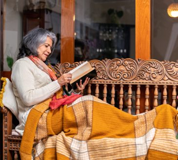 Cozy scene of a senior woman relaxing on a wooden bench with a blanket, reading a book and enjoying the comfort of her home