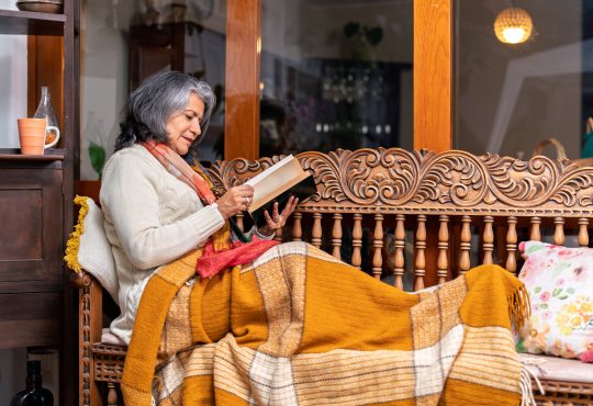 Cozy scene of a senior woman relaxing on a wooden bench with a blanket, reading a book and enjoying the comfort of her home