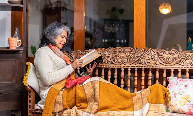 Cozy scene of a senior woman relaxing on a wooden bench with a blanket, reading a book and enjoying the comfort of her home