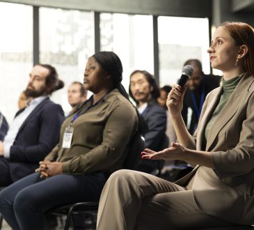 Young adult businesswoman in audience interacting with presenter, asking questions and engaging in debate. Mixed age participants include managers, employees and investors at summit.