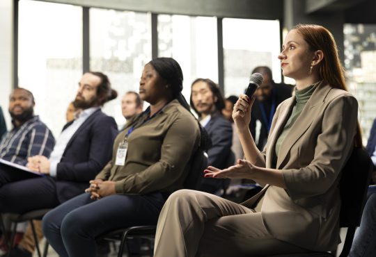 Young adult businesswoman in audience interacting with presenter, asking questions and engaging in debate. Mixed age participants include managers, employees and investors at summit.