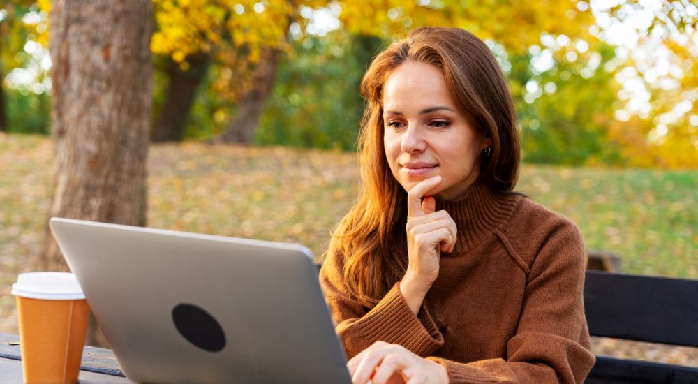 A mid-adult woman watching online webinar and studying on laptop while sitting in autumn park. Adult education and online reskilling.