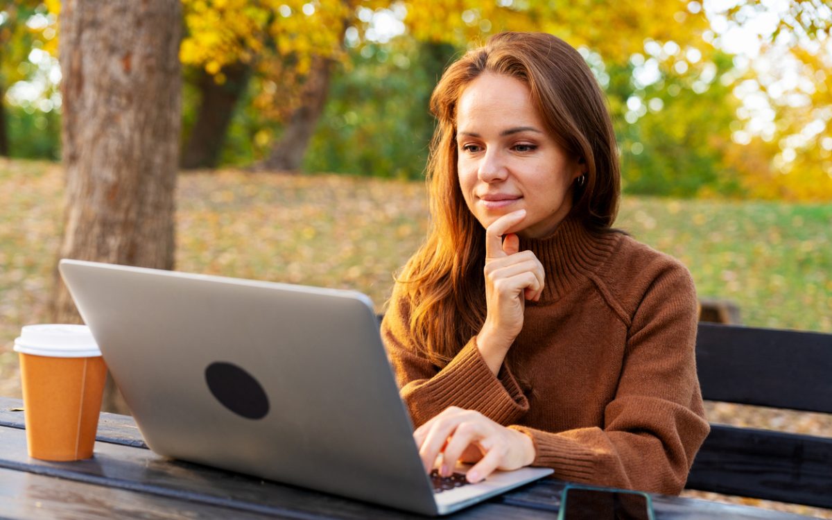 A mid-adult woman watching online webinar and studying on laptop while sitting in autumn park. Adult education and online reskilling.