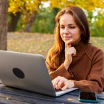 A mid-adult woman watching online webinar and studying on laptop while sitting in autumn park. Adult education and online reskilling.