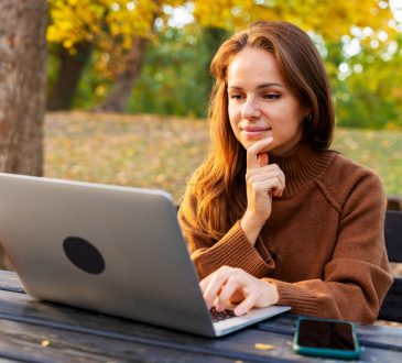 A mid-adult woman watching online webinar and studying on laptop while sitting in autumn park. Adult education and online reskilling.