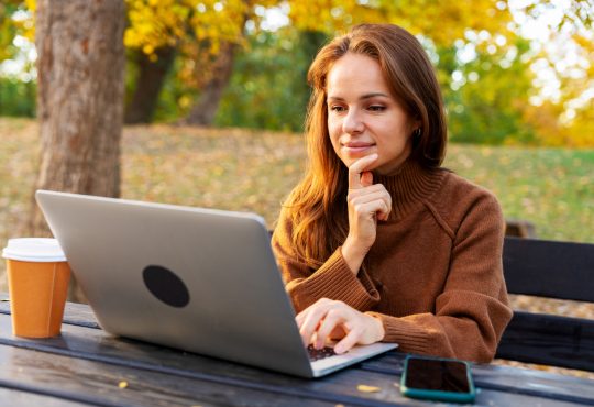 A mid-adult woman watching online webinar and studying on laptop while sitting in autumn park. Adult education and online reskilling.