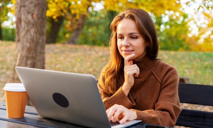 A mid-adult woman watching online webinar and studying on laptop while sitting in autumn park. Adult education and online reskilling.