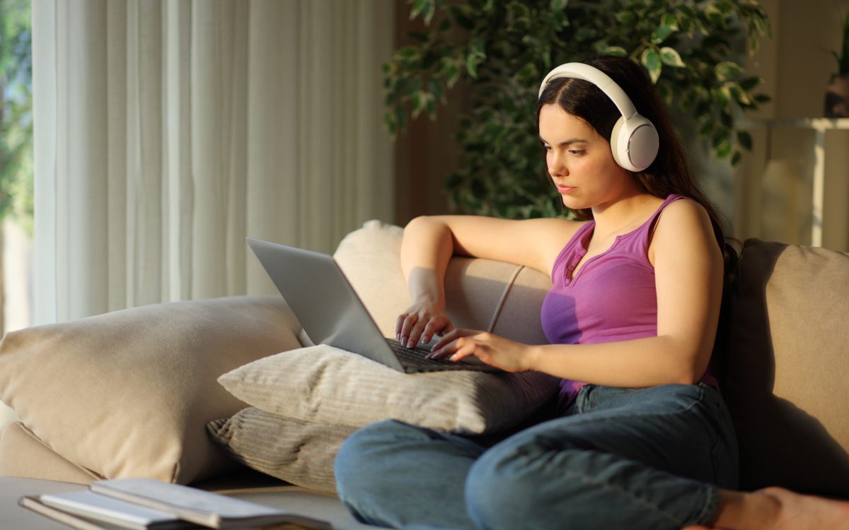 Student wearing headphone typing on laptop e-learning sitting on a couch at home