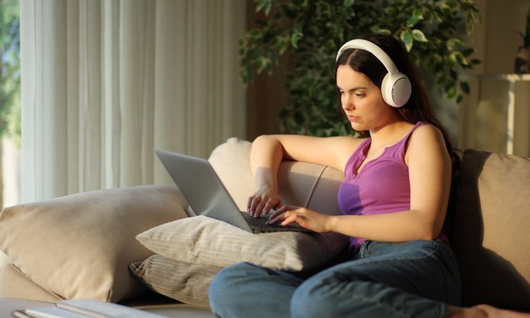 Student wearing headphone typing on laptop e-learning sitting on a couch at home